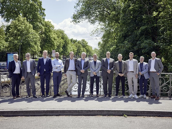 A group of twelve men in formal attire stands together on a bridge, surrounded by lush green trees and a bright blue sky.