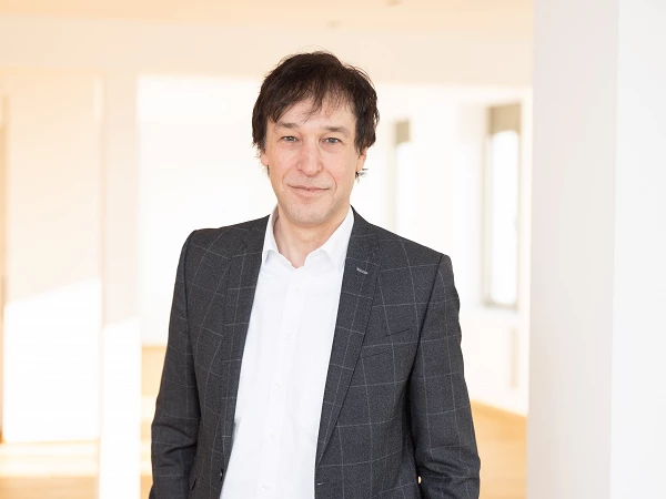 A man with dark hair and a slight smile stands in a well-lit room with neutral walls and wooden flooring, wearing a gray patterned blazer over a white shirt.