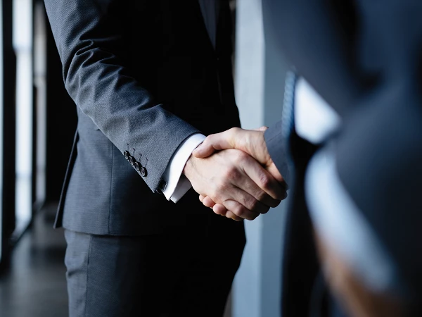 Two men in suits are shaking hands in a modern setting with large windows in the background.