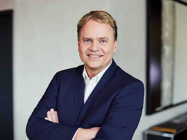 A smiling man in a navy blazer stands with arms crossed in a modern office setting.