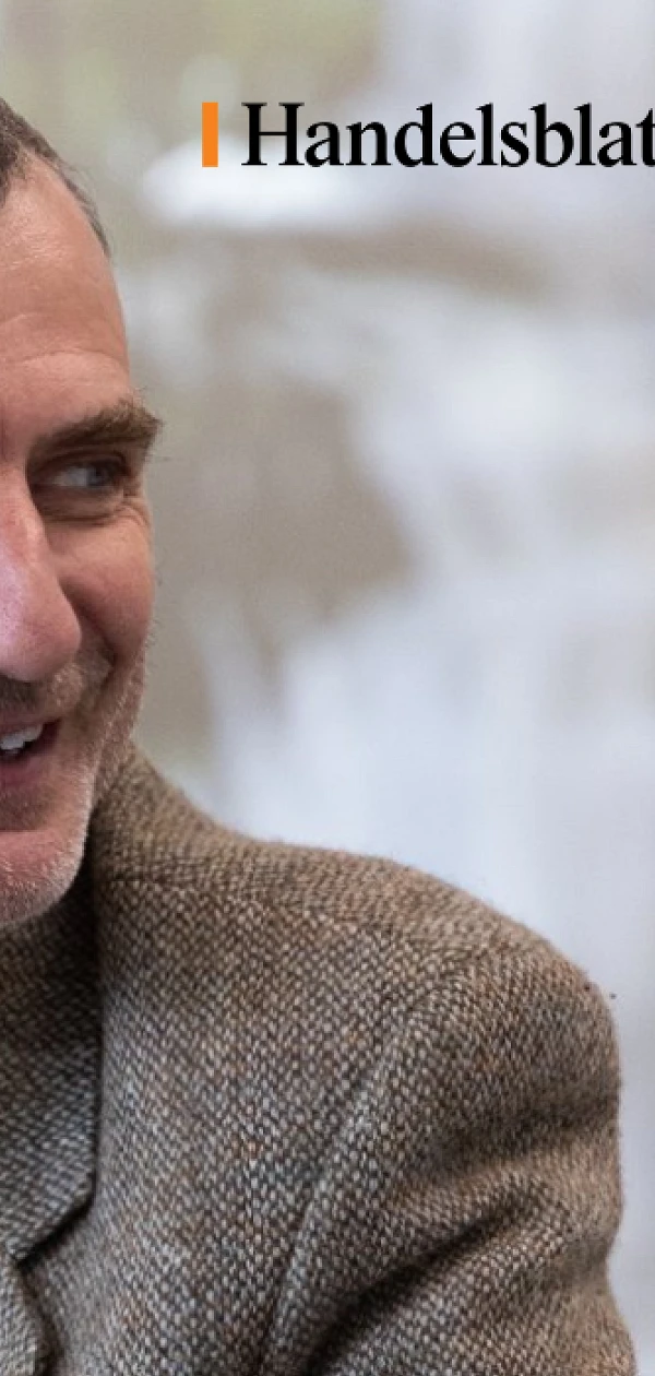 Ein lächelnder Mann in einem grauen Jackett sitzt an einem Tisch und blickt zur Seite. Im Hintergrund sind unscharfe Personen und eine helle Umgebung sichtbar. In der oberen rechten Ecke befindet sich das Logo von Handelsblatt.