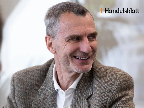 Ein lächelnder Mann in einem grauen Jackett sitzt an einem Tisch und blickt zur Seite. Im Hintergrund sind unscharfe Personen und eine helle Umgebung sichtbar. In der oberen rechten Ecke befindet sich das Logo von Handelsblatt.