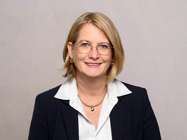 Frau mit blonder Bob-Frisur und Brille im Business-Outfit, lächelnd vor hellem Hintergrund