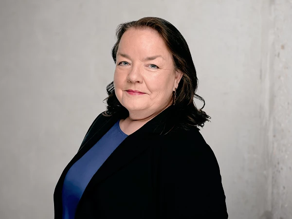 Frau mit dunklen Haaren im schwarzen Blazer und blauem Oberteil vor grauer Wand