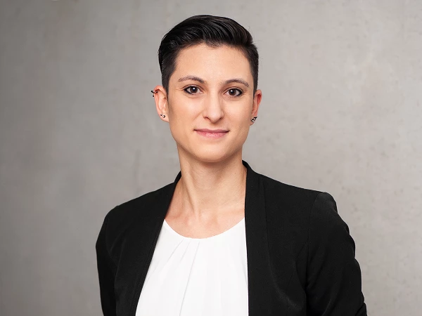 Frau mit kurzem dunklen Haar im schwarzen Blazer und weißer Bluse vor Betonwand