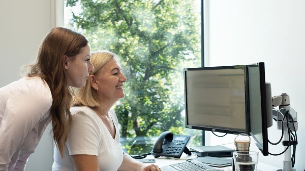 Zwei Kolleginnen blicken gemeinsam auf einen Bildschirm, helle Büroatmosphäre
