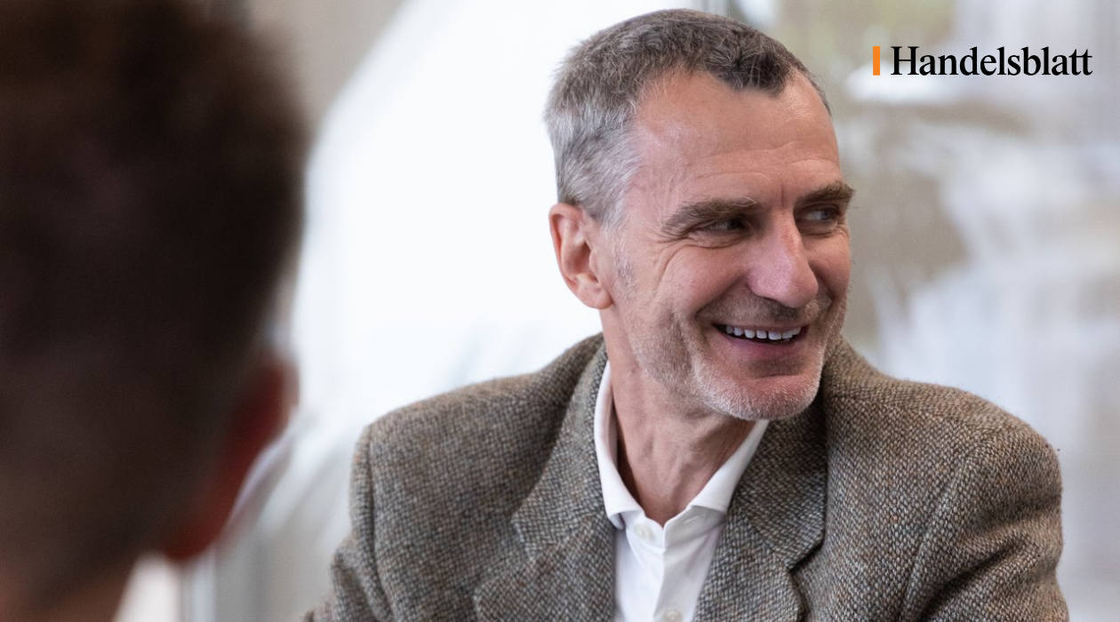 Ein lächelnder Mann in einem grauen Jackett sitzt an einem Tisch und blickt zur Seite. Im Hintergrund sind unscharfe Personen und eine helle Umgebung sichtbar. In der oberen rechten Ecke befindet sich das Logo von Handelsblatt.