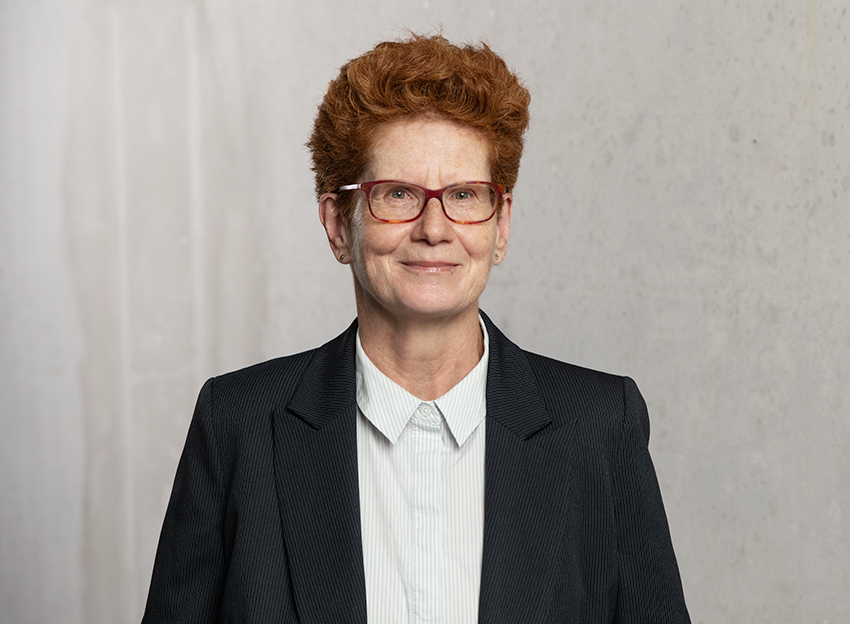 Frau mit roten Haaren und Brille im Business-Outfit vor neutralem Hintergrund